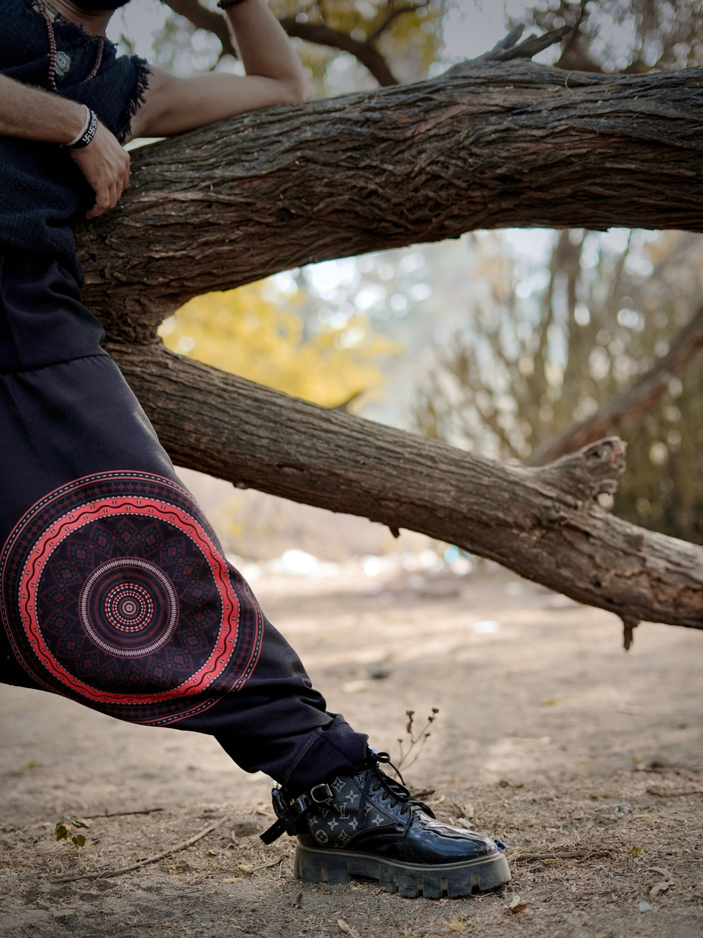Person wearing dark pants with a red and purple circular design, standing next to a tree in a natural setting.
